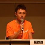 オレンジ色のシャツを着た三浦さんが右手でマイクを持ち話をしている様子をアップで撮影した写真