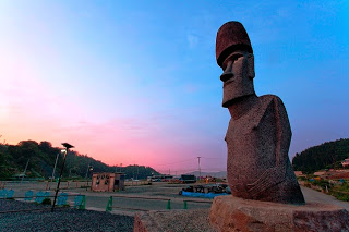オレンジ色に染まっている朝焼けの空と木々が立ち並ぶ林を背景に、周囲にバリケードが設置された工事中の中に立つ石造りのモアイ像が写された写真