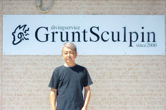 グレーの短髪で黒いTシャツを着た男性が、レンガ模様の壁に取り付けられた「diving service GruntSculpin since 2000」と書かれた白い看板の前に立っている写真