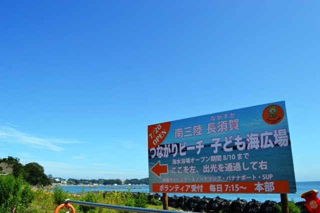 海と青空を背景に、「つながりビーチ子ども海広場」と書かれたカラフルな看板が立っており、オープン日や案内が詳しく書かれている案内看板の写真