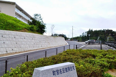 白いコンクリートの擁壁が緩やかな傾斜のある道路沿いに続き、緑の植栽と集合住宅が見える町営名足復興住宅の入口付近の写真