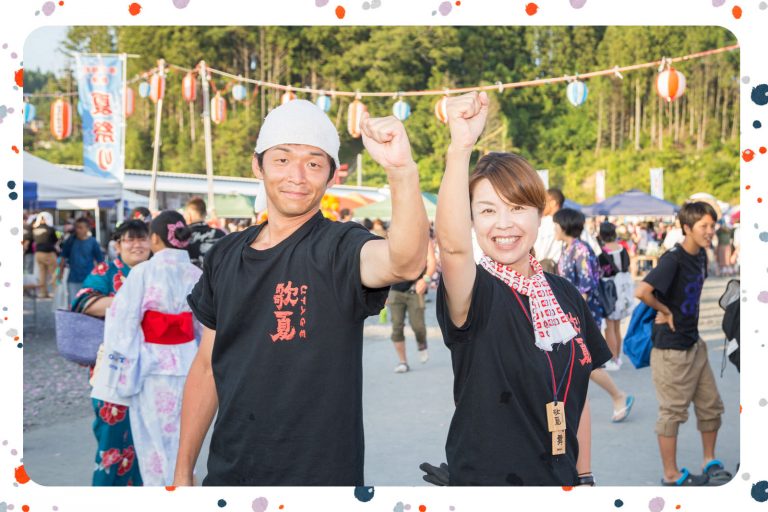 背景には浴衣姿の人々や提灯が並ぶ賑やかな夏祭り会場で、お揃いの黒いシャツを着た二人の男女が腕を高く突き上げて笑顔を見せている写真