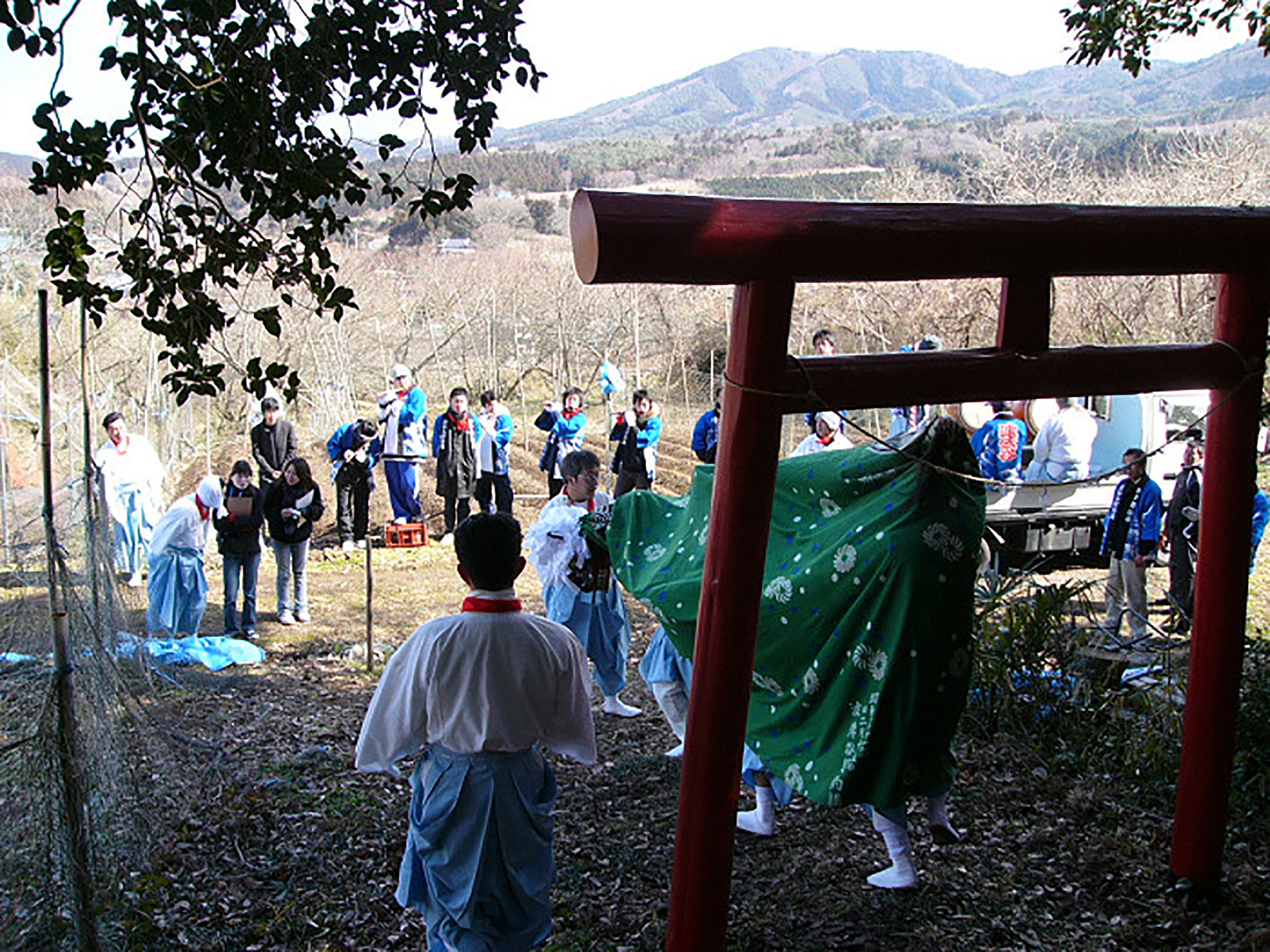 山間の神社の鳥居前で獅子舞と白装束の人々が儀式を行い、周囲で法被姿の数名が楽器を演奏している写真