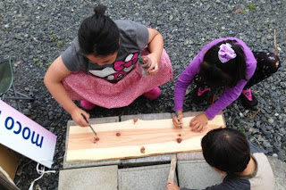 屋外の砂利の上で三人の子どもが木の板に向かって作業している様子を写しており、うち二人は工具を使って釘を打ち込んでいて、もう一人はその様子を見守っており、左側には「MOCO」と書かれた看板が部分的に見えている写真