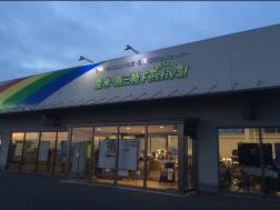 夕暮れ時で空がやや暗く、建物の正面には虹色のラインが描かれ、緑色の文字で「登米・南三陸フェスティバル」と書かれている店舗の外観の写真