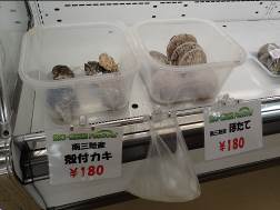 棚に並べられた2つのプラスチック容器に、殻付きの商品が入っており、それぞれに「殻付カキ180円」と「ほたて180円」と書かれたポップが貼られている様子の写真