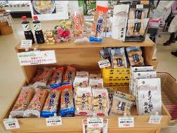 木製の陳列棚に、さまざまな種類の食品がパッケージされた状態で並べられており、左上には2本の醤油のボトルが置かれている様子の写真