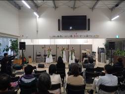 壁には大画面のモニターが設置され、スピーカーなどの音響機器、天井が高く梁が見える室内で、ステージに向かって複数の人が座って見ているイベント会場の様子で、ステージには白いワンピースを着た3人の女性が踊っている様子の写真