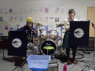 壁にさまざまな絵や作品が飾られた屋内で、ドラムセットの左横に座った男性がギターを弾き、右手前に立った女性が鍵盤ハーモニカを演奏している写真