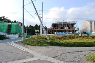 電柱が立つ舗装道路のそばで木造住宅が骨組みの状態で建設されており、その奥には足場とシートに覆われた建物も見える写真