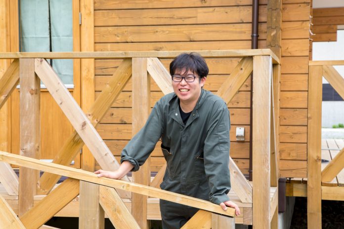 木製の建物前のスロープで、灰色の作業着を着た黒縁メガネの男性が木製手すりに手をかけて笑顔で立っている写真