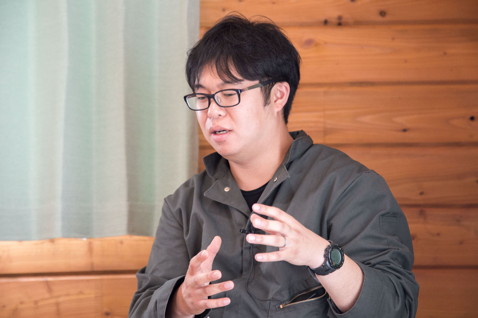木の壁とレースのカーテンがかけられた室内で、灰色の作業着を着た黒縁メガネの男性が身振り手振りで話しをしている写真