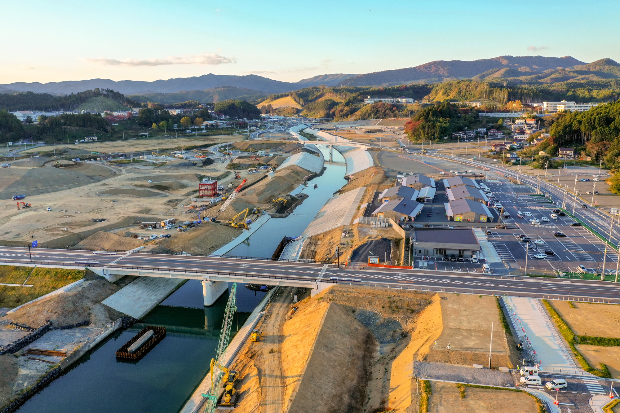 夕方の柔らかい光の中で山に囲まれた広い土地にかかる橋の上から撮影され、橋の下には細くて直線的な川が流れ、両岸で工事や整備が行われている様子が見られ、右側には整然とした建物群と駐車場が広がり、背景には山々が連なっている写真