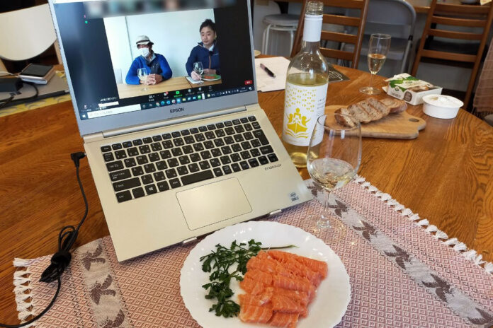 画面に2人の男性が映し出されているノートパソコンでZoom会議をしながら、テーブルの上に白ワインと銀鮭のコンフィが盛り付けられた白い皿、グラスやパンが並べられている写真