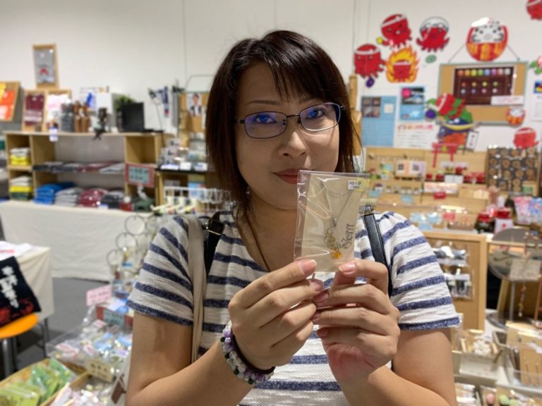 女性が土産物店のような売り場で、小さなパッケージに入ったクセサリーの商品を笑顔でカメラに向けて差し出している写真