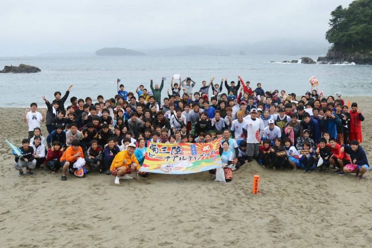 青い海と白い砂浜を背景に、「南三陸ビーチアスティメット」の横断幕を中央の方々が持っており、大勢の参加者が集まった集合写真