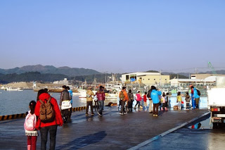 港の岸壁に親子連れやグループが集まり、漁船や港の建物を散策している様子の写真