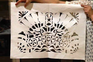 幾何学模様や花のようなデザインが施された大判の切り絵の作品を、レース柄の服を着た人物が正面で持ち上げて見せている写真