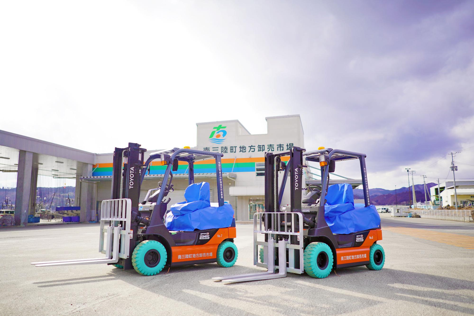 「南三陸町地方卸売市場」と書かれた市場を背景に電動フォークリフトが2台横並びに停車している写真
