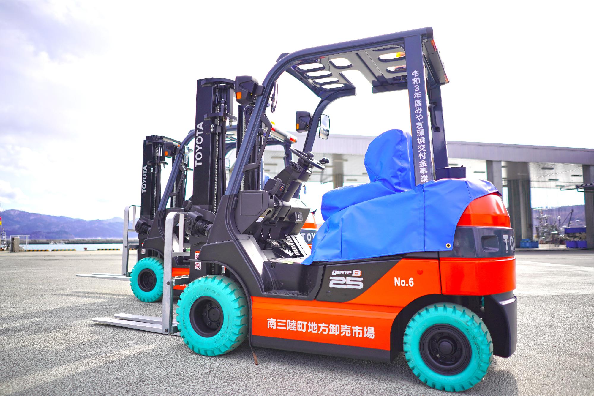 青いカバーのかけられた運転席とミントグリーンのタイヤが特徴的な電動フォークリフトが、広い屋外ヤードに停まっている写真