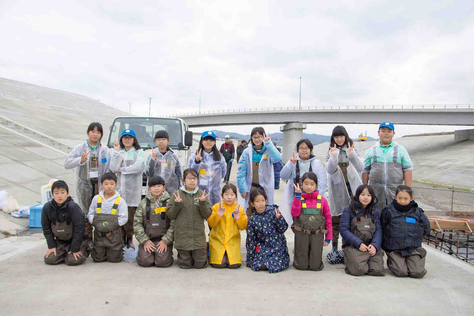 奥に橋がかかった堤防で川の活動に参加する子どもたちが2列に並んだ集合写真