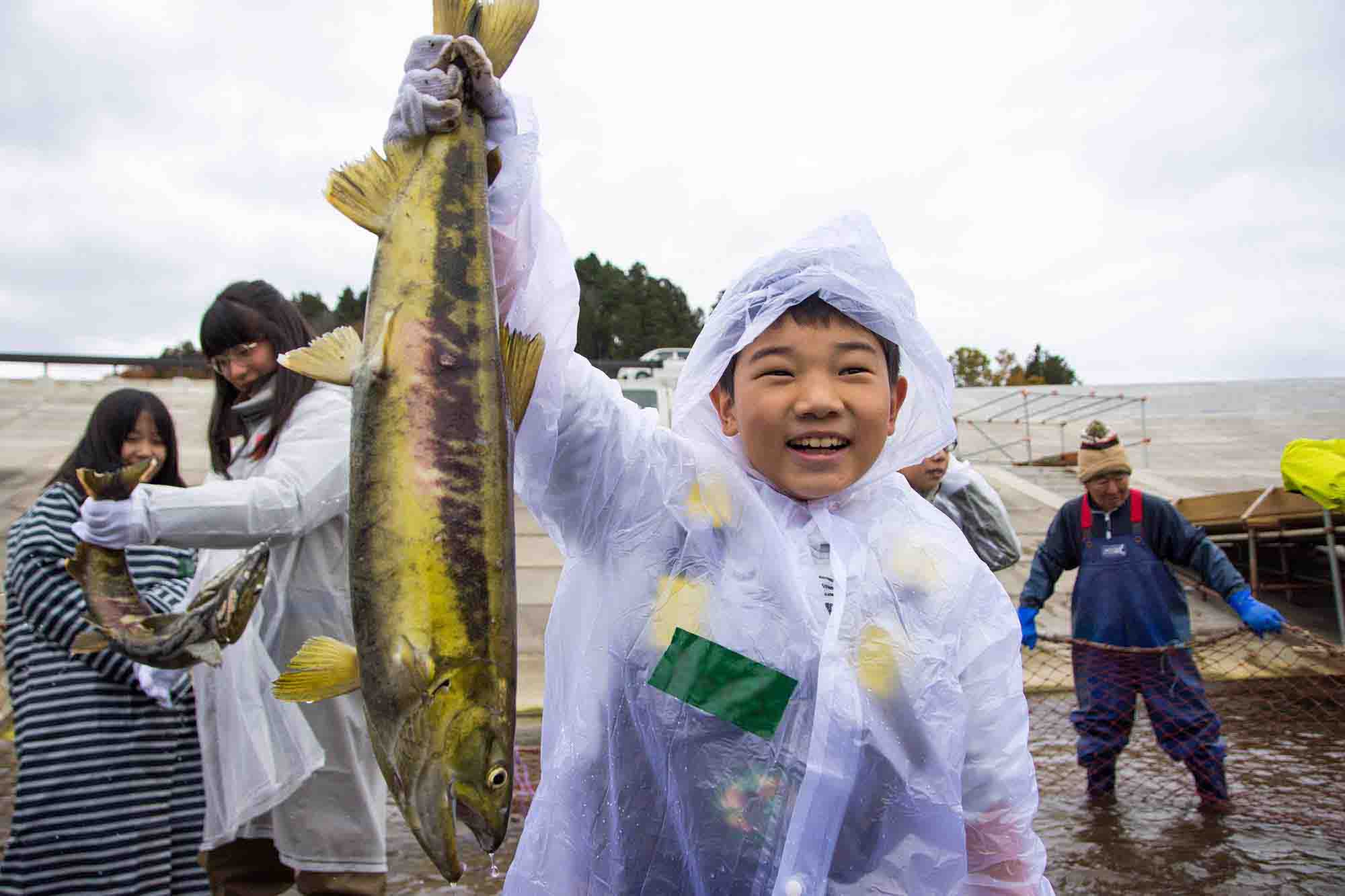 レインコートを着た男の子が大きな鮭を持ち上げた笑顔の写真
