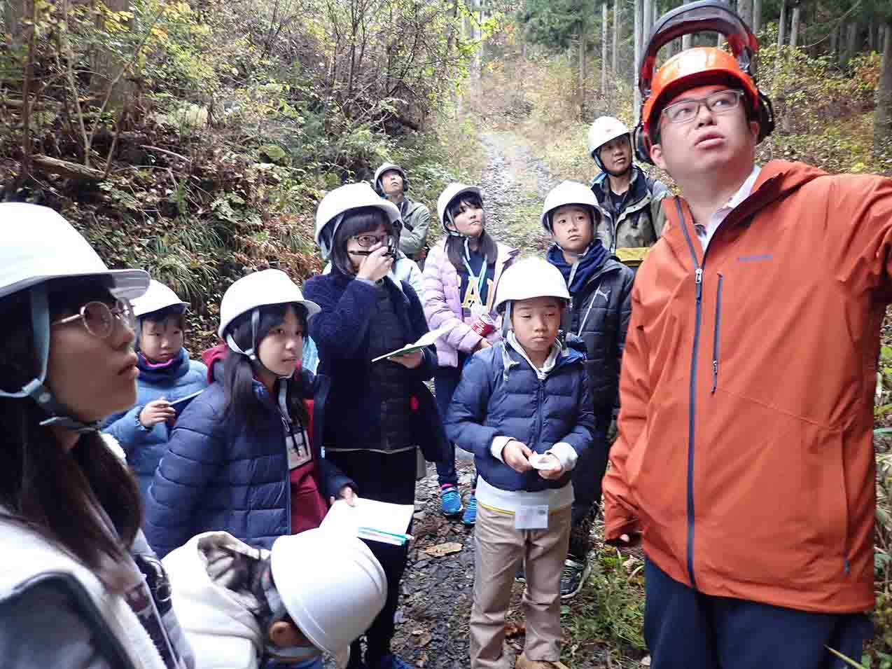 森の中でヘルメットをかぶった子どもたちがガイドの話を聞いている様子の写真