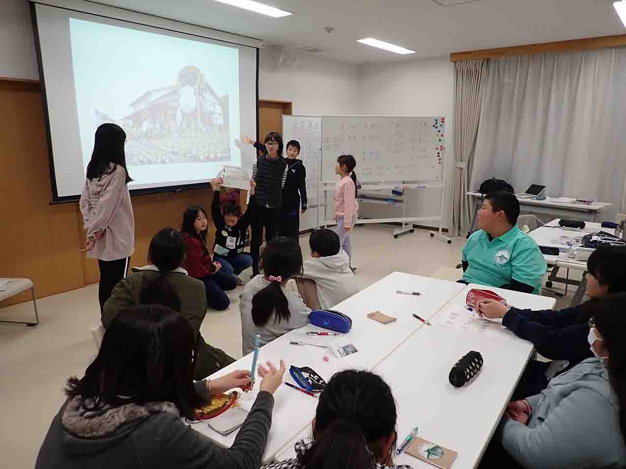 屋内で映像が映し出されたスクリーンの前に立つ子どもたちが発表している様子の写真