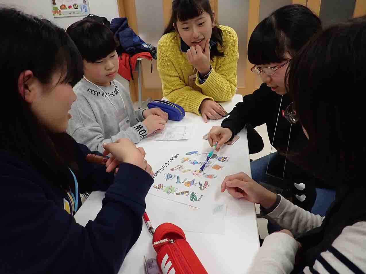 5人の女の子たちがテーブルの上に置かれたイラストが描かれた紙を見て話し合っている様子の写真