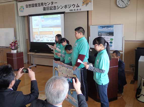 前列に座っている2人の男性がカメラを向けているなか、お揃いの緑色のポロシャツを着た5人の隊員の子どもたちが前方に横一列に並び発表を行っている様子の写真