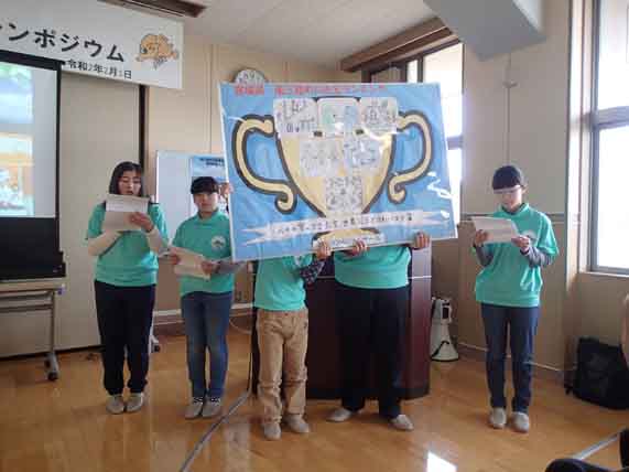 中央に立つ2人が優勝カップの様なイラストが描かれた大きなボードを持ち、左側の女の子が紙を持って発表を行っている様子の写真