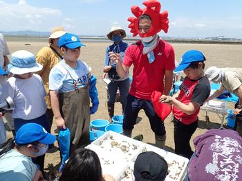 干潟でカニの帽子をかぶった男性が子どもたちに生き物を見せている写真