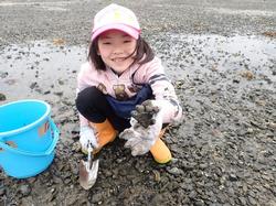干潟にしゃがんでスコップを持ち、採取した生き物を手に笑顔を見せる女の子の写真