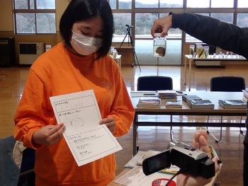 右側で標本瓶を持った手の傍に立つ女の子がカメラに向かって観察記録を見せている写真