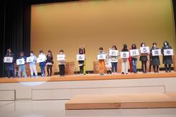 舞台の上で1人1枚の文字カードを持ち、全員で並んでスローガンを掲げている発表の写真