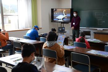 教室の黒板の左側に設置された大型モニターの傍に立つ男性の話を、自席に座って熱心に聞き白鳥について学んでいる子どもたちの写真