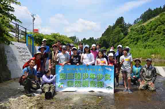 川の中で「南三陸少年少女自然調査隊」と書かれた横断幕を持ち集合写真を撮る子どもたちの写真