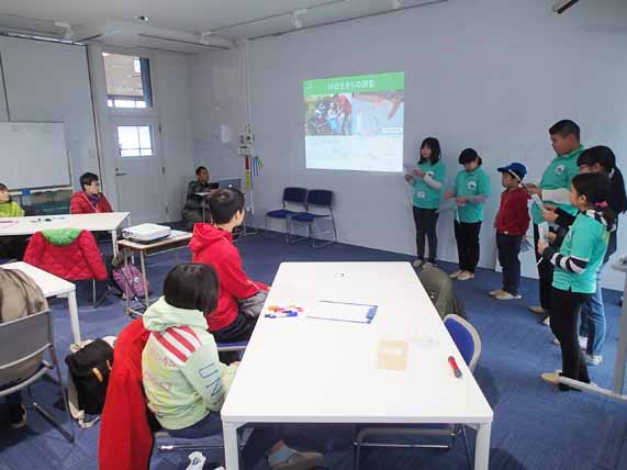 参加者が見ているなか、資料が映し出された教室の壁の前に立ち発表している子どもたちの写真