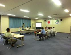 前方にはスクリーンと日本の国旗、市旗が掲示された会議室で、10人ほどの参加者がコの字型に並べられた机の周りに座り会議が行われている様子の写真