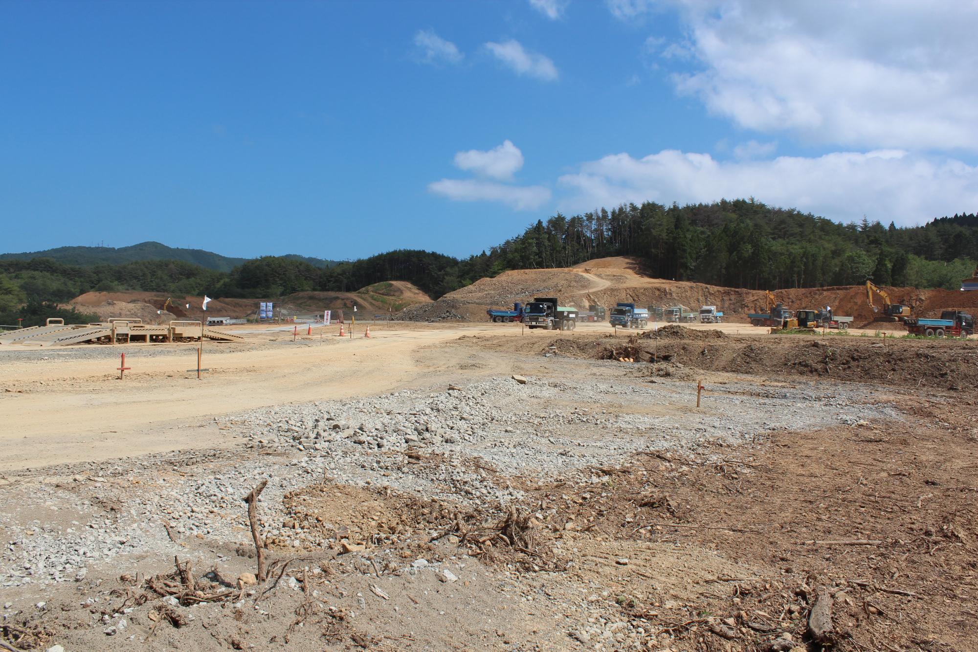 山あいの開けた土地で、重機やトラックが多数並び造成作業を進めている様子を地上から写した、青空の下の造成工事中の写真。
