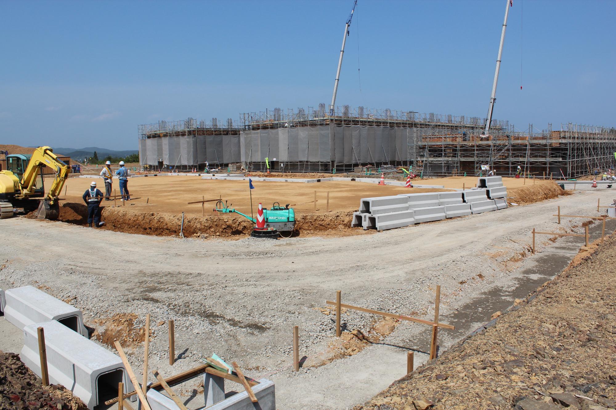 重機や作業員がいる中、整地された広い土地に基礎工事が進められており、奥には足場とネットに覆われた建物が建設中の様子が見える、災害公営戸倉住宅を作っている写真