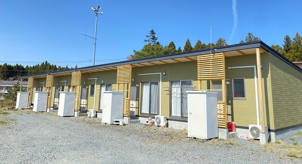 複数戸が連なった薄緑色の平屋建て仮設住宅で、各戸の前にエアコン室外機や収納が設置されている峰畑定住促進住宅の外観写真
