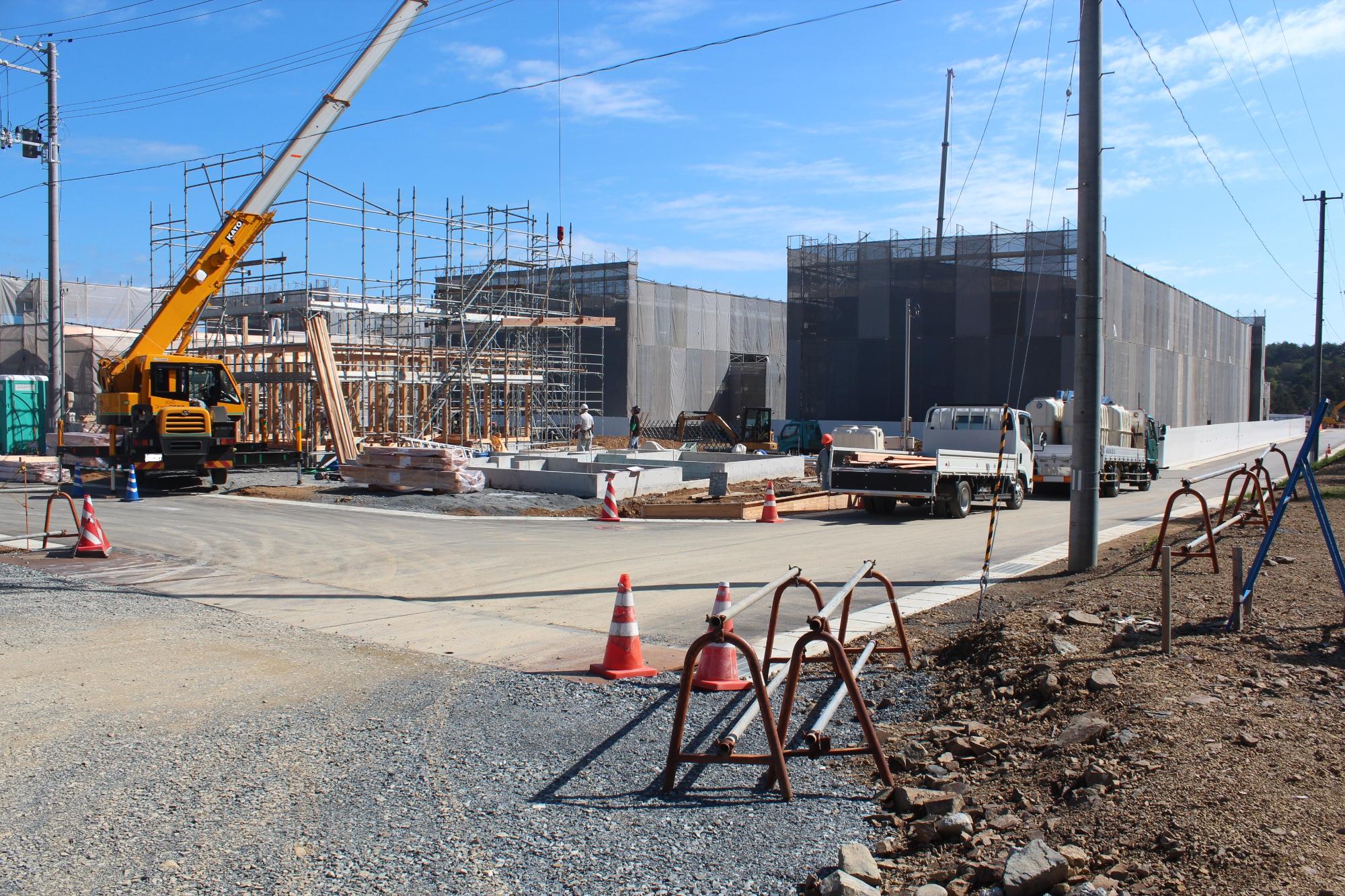 大型クレーンが稼働し、建物の骨組みが組み立てられている現場の奥に、灰色のネットに覆われた大きな建物が並ぶ、工事が進行中の災害公営戸倉住宅を作っている写真