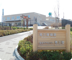 植栽に「南三陸病院」の文字がデザインされた木製の看板があり、その横の緩やかな上り坂の上に南三陸病院がたっている写真