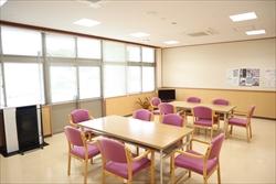 机に6脚ずつピンクの椅子が設置され、奥にはテレビが見えるフローリングの室内写真