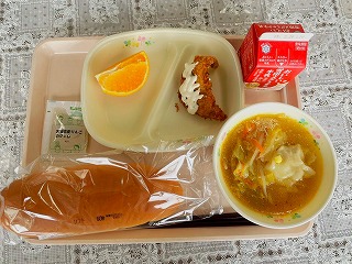ピンクのトレーに給食が左から時計回りにりんごジャム、カットされたデコポン、手作りたらフライ、牛乳、みそワンタンスープ、ソフトパンが並んでいる写真