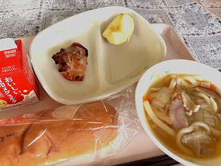 ピンクのトレーに給食が左から時計回りに牛乳、鶏肉のマーマレード焼き、りんご、ちゃんぽんうどん、米粉パンが並んでいる写真