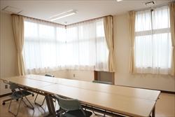 長机が2台くっつけられ、角部屋の窓にはカーテンがある明るい会議室内の写真