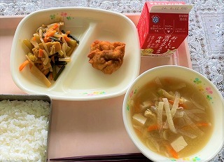 ピンクのトレーに給食が左から時計回りにたけのこと山菜の煮物、鶏肉の唐揚げ、牛乳、ひきな汁、ごはんが並んでいる写真