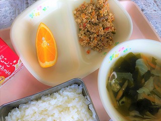 ピンクのトレーに給食が左から時計回りに牛乳、オレンジ、ガパオライスの具、おくらとわかめのスープ、ごはんと並んでいる写真